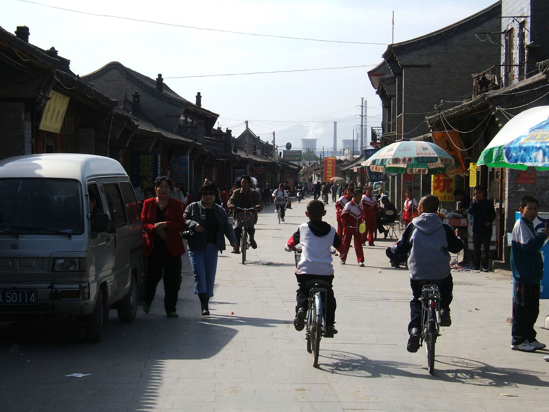 Dated busy old-city street with low buildings, bicycles, pedestrians and distant stacks