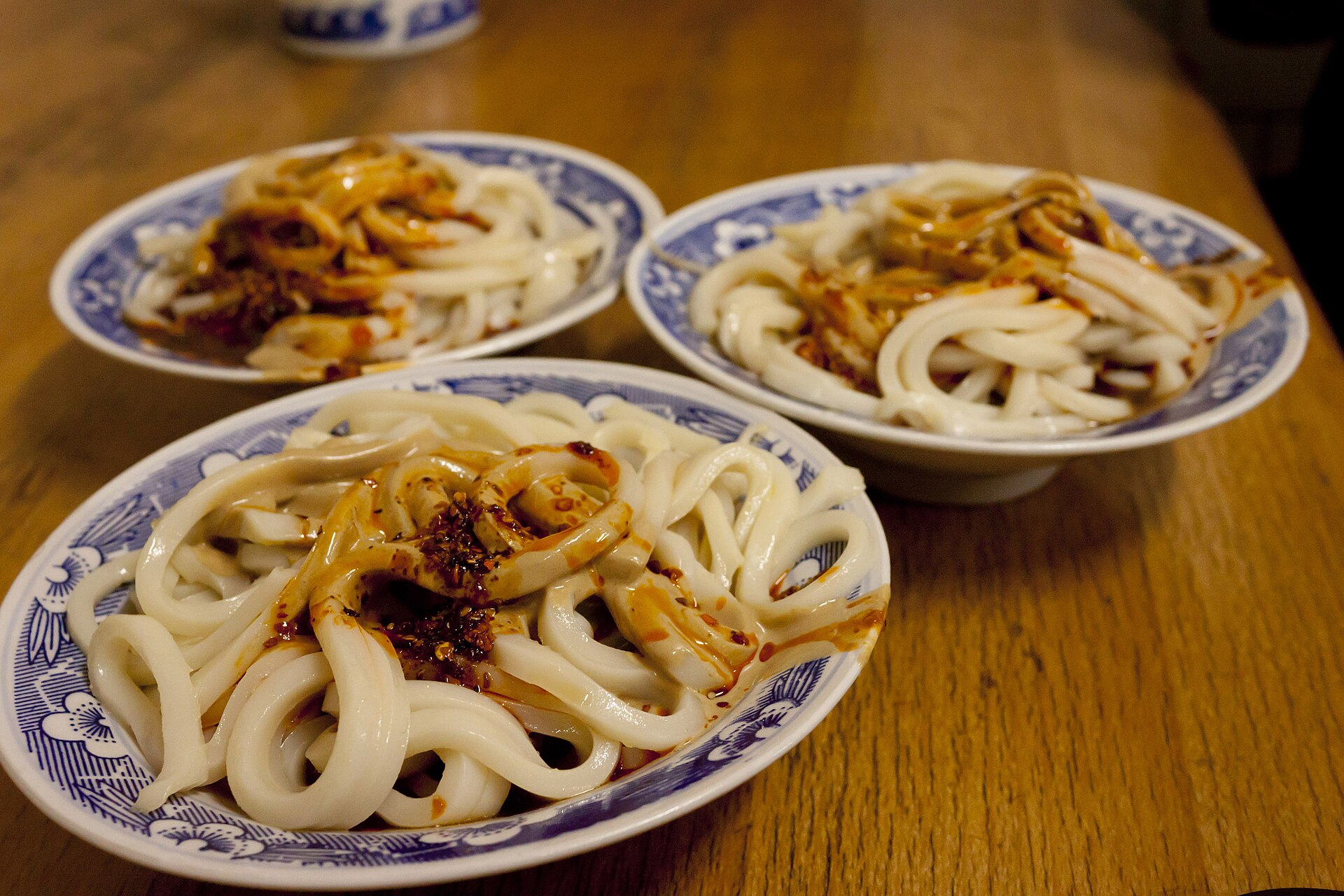Three bowls of pale liangpi strips with dark sauce and chili oil